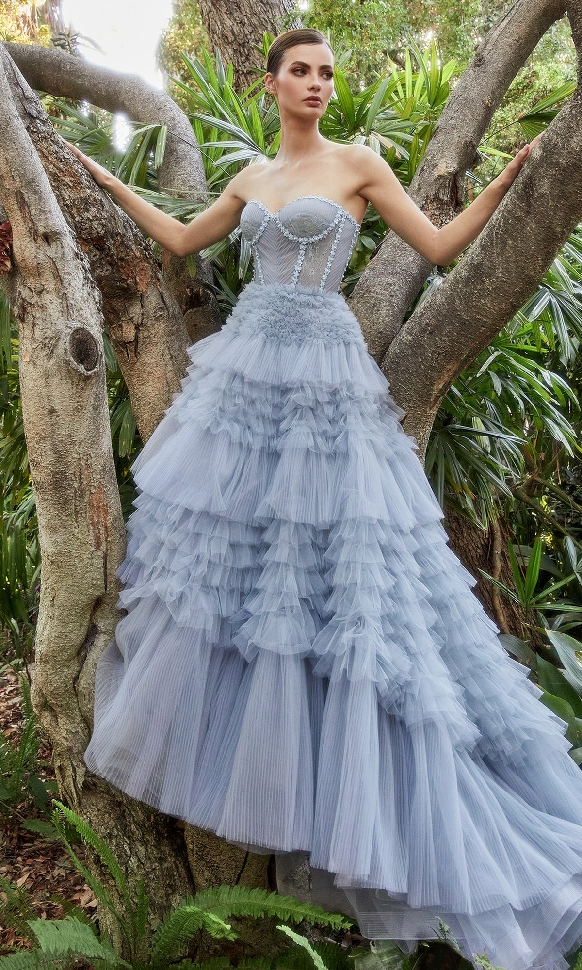 A woman wearing the Strapless Tiered Long Prom Ball Gown A1017 in light blue stands outdoors among tree branches and lush greenery, with delicate ruffles accentuating her look.